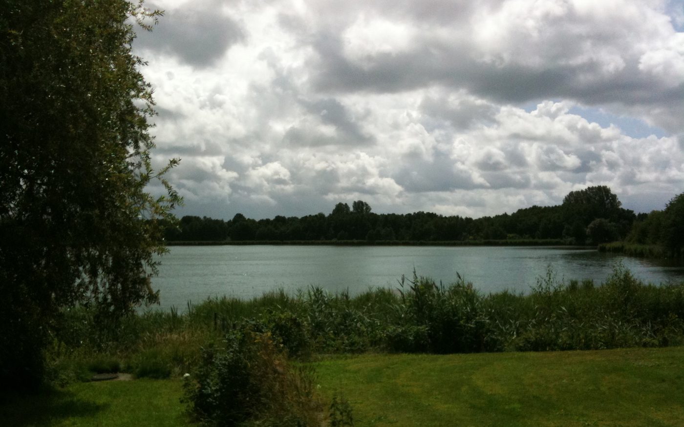 Der Fockbeker See unter Wolken, von einem Wiesenufer aus gesehen